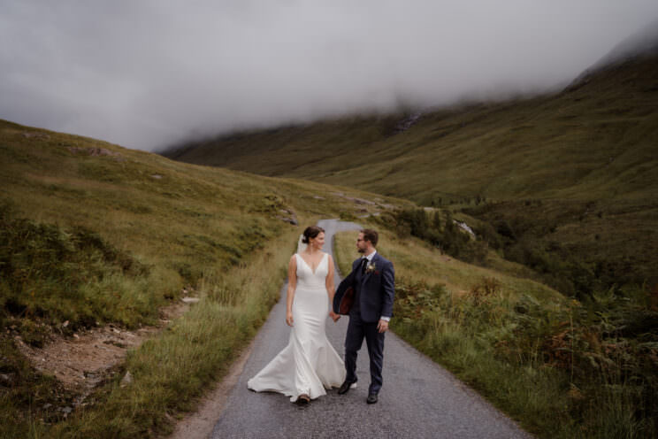 A newlywed couple walk hand in hand down a narrow road during their Scotland elopement.