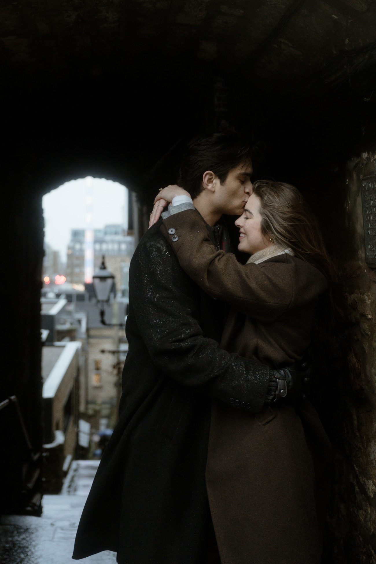 A couple embraces in a narrow, historic stone alleyway in Edinburgh. The man, dressed in a dark coat, gently kisses the woman on the forehead as she smiles and wraps her arms around him