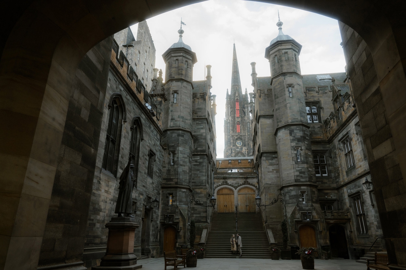 A couple stands at the base of a grand stone staircase in a historic courtyard, framed by towering gothic-style architecture of Edinburgh New College