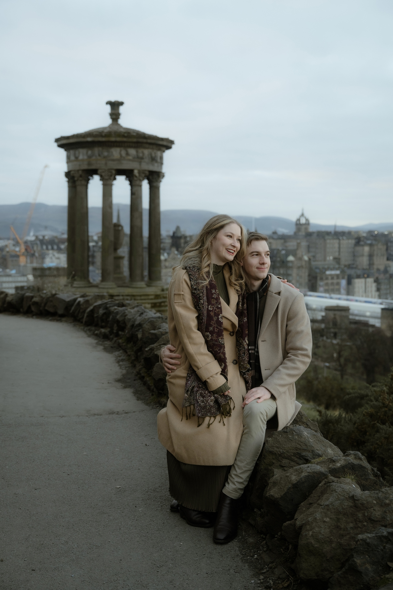 A couple sits on a stone ledge on Calton Hill, overlooking the city of Edinburgh. The woman, dressed in a long beige coat and a patterned scarf, smiles warmly while leaning into her partner, who sits beside her with his arm wrapped around her