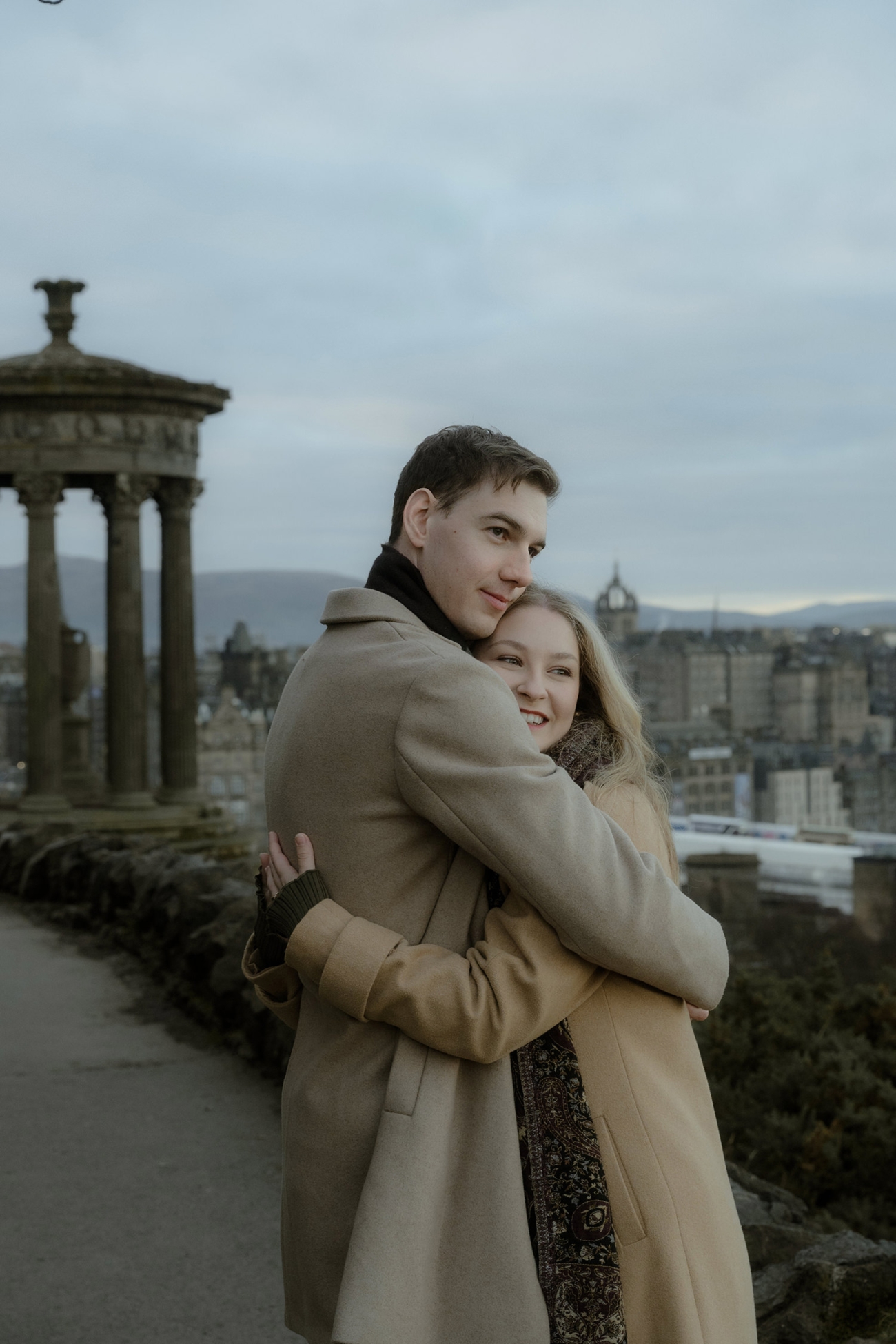 The man, wearing a beige coat and a black scarf, gazes into the distance while the woman, also in a beige coat, smiles and leans into his embrace. The historic Dugald Stewart Monument in the background