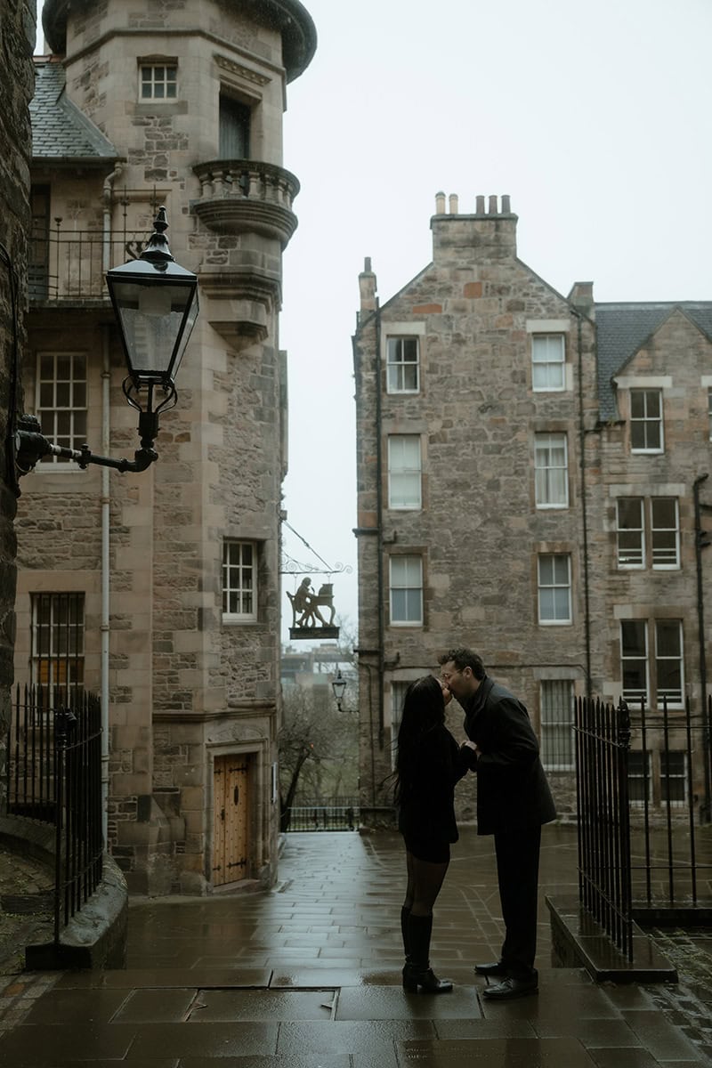 A couple share a kiss in the rain at Lady Stair's Close, Edinburgh, surrounded by historic stone tenements and a Victorian gas lamp, with the Writer's Museum turret in the background