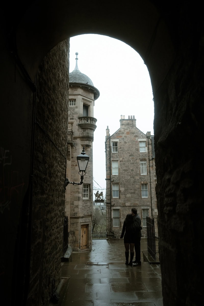 A couple stand silhouetted in a stone archway at Lady Stair's Close in Edinburgh, looking out towards the Writer's Museum courtyard on a rainy day