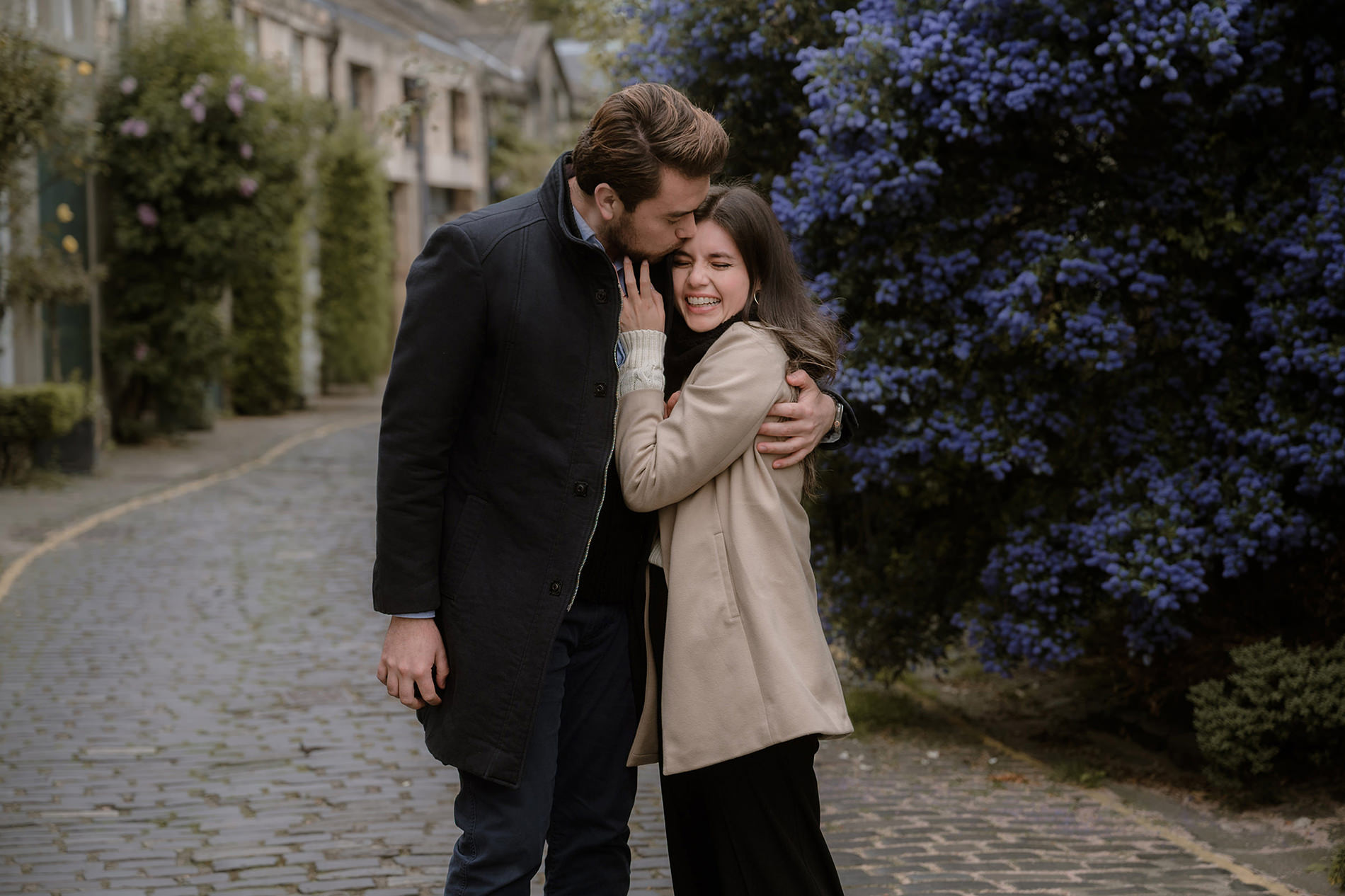 Post-proposal moment - couple embracing on a quiet cobblestone street lined with stone houses and lush greenery. The man in a dark coat kisses the woman’s forehead as she smiles with her eyes closed, standing beside a large bush of vibrant purple flowers on Circus Lane