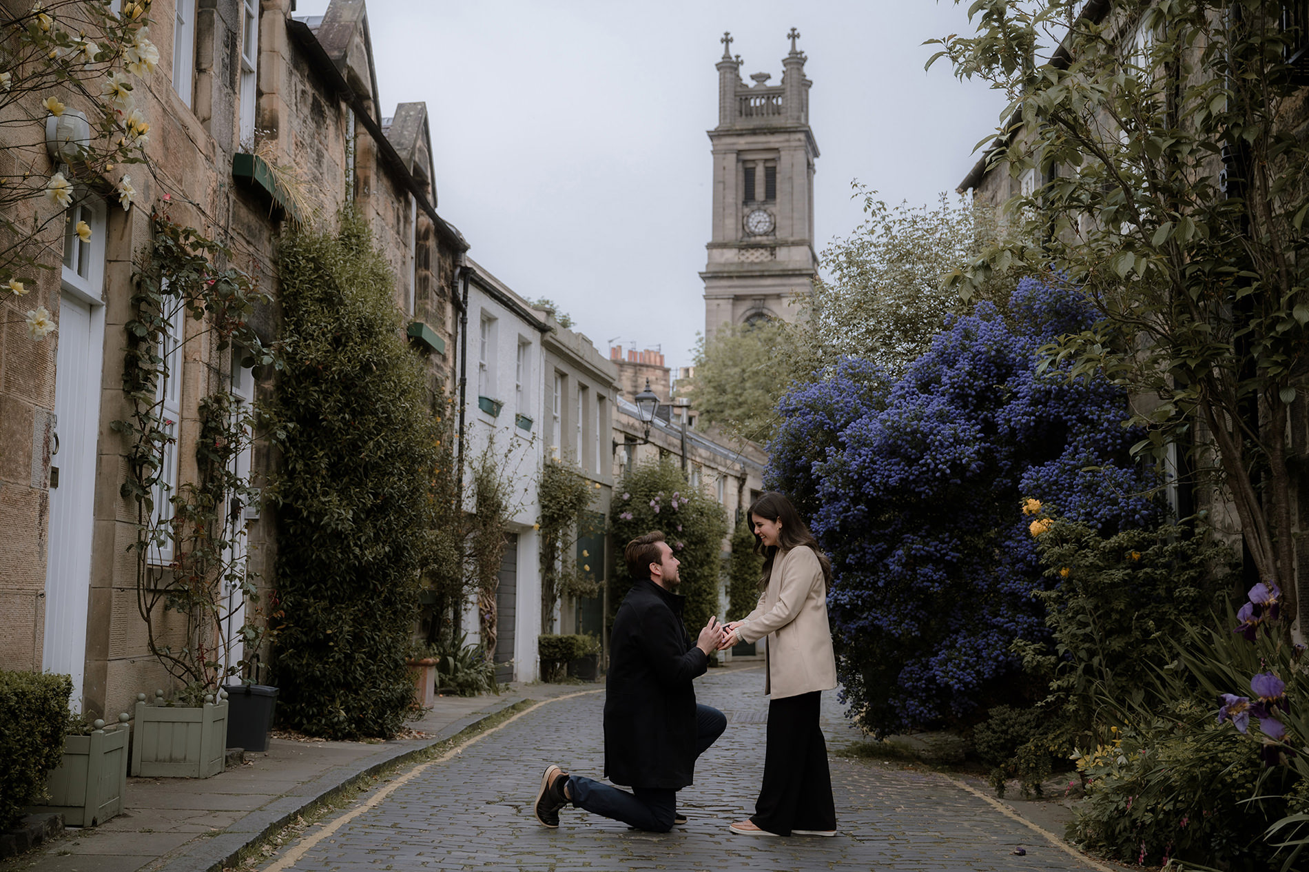 Proposal in Edinburgh - on a charming cobblestone Circus Lane with ivy-covered houses and a church tower in the background, a man kneels on one knee holding a woman’s hands in a proposal. She stands facing him in a light coat and long dark skirt, framed by colorful flowers and spring greenery