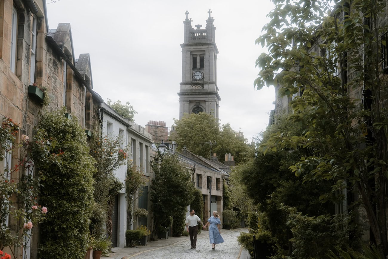 A couple walk hand in hand along the cobblestones of Circus Lane in Edinburgh's New Town, surrounded by ivy-covered mews houses, climbing roses and the clock tower of St Stephen's Church rising behind them