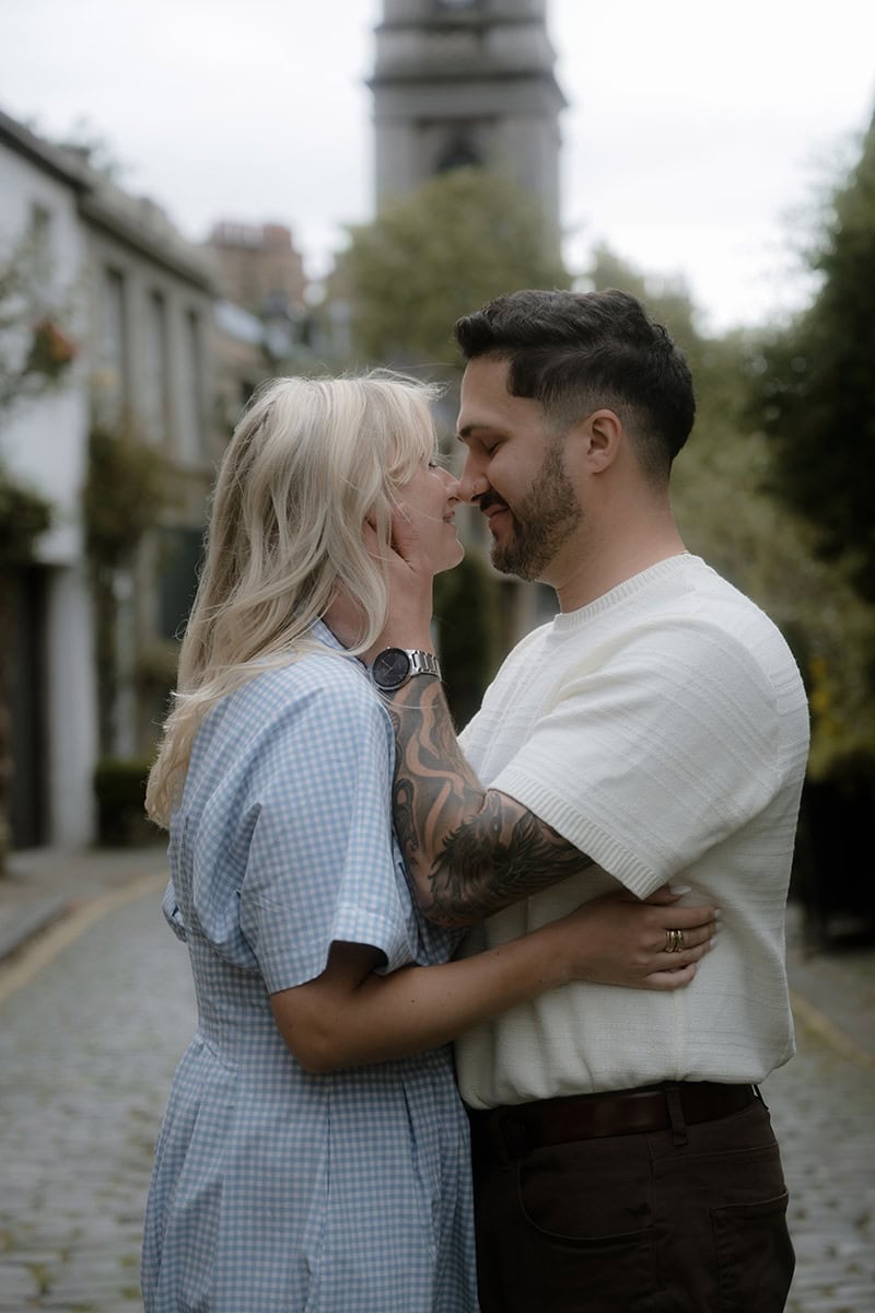 A couple share an intimate moment on Circus Lane in Edinburgh, foreheads almost touching and smiling, with the cobblestone mews and St Stephen's Church tower softly blurred behind them