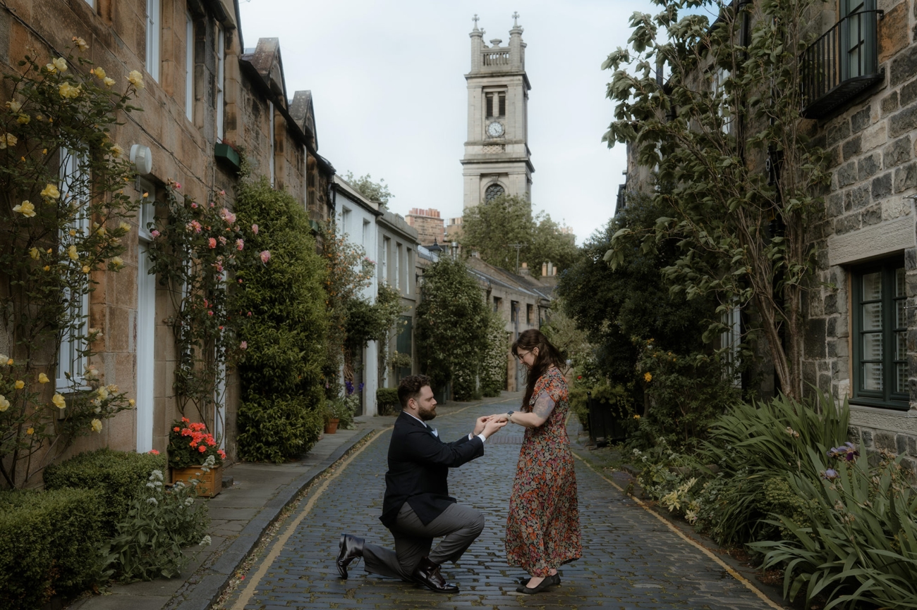 Man dressed in a suit gets down on one knee at Edinburgh's Circus Lane when proposing to his girlfriend in the spring, surrounded by greenery, flowers and with a towering cathedral clock tower in the background
