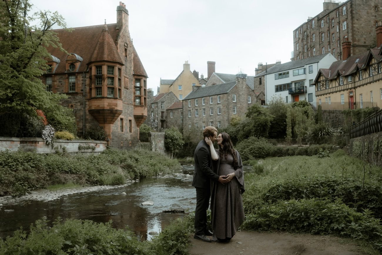 A couple share a kiss beside the Water of Leith in Dean Village, Edinburgh, with the Victorian Well Court building and historic stone tenements reflected in the river behind them