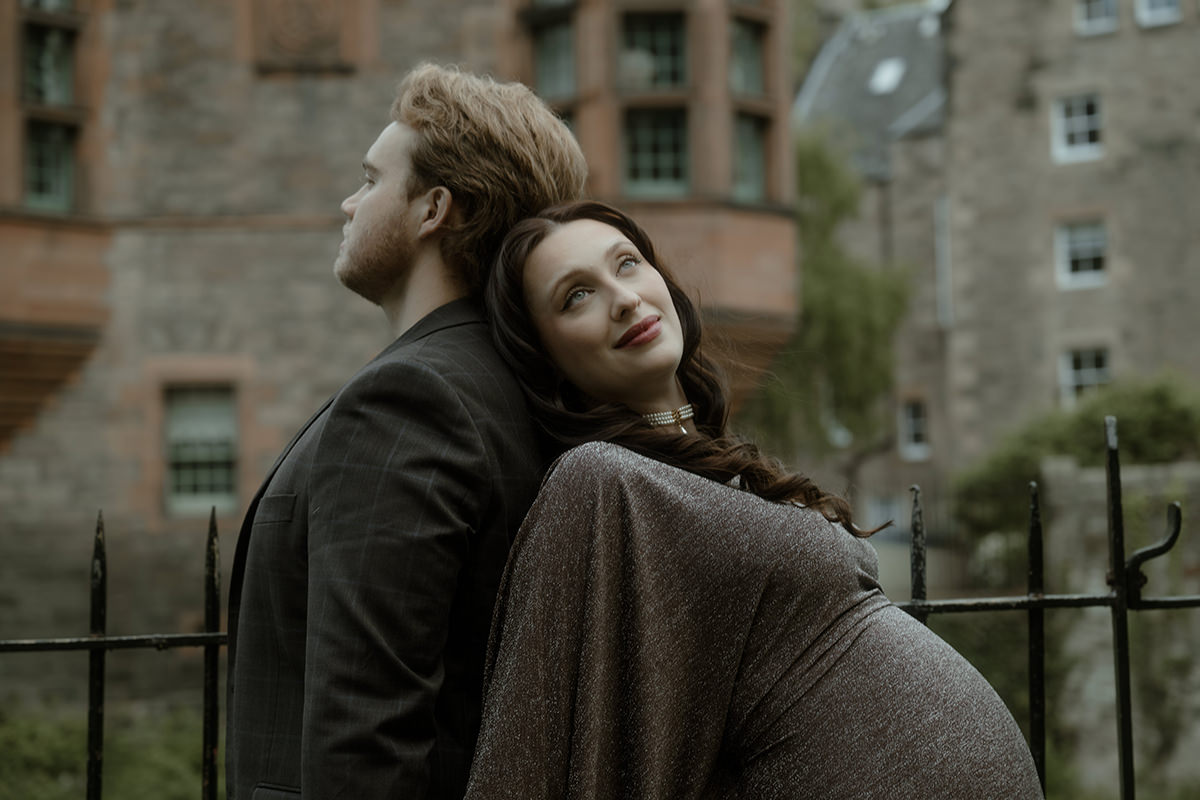 A woman in a grey maxi dress leans back against her partner during an engagement session in Dean Village, Edinburgh, historic stone tenements softly blurred behind them