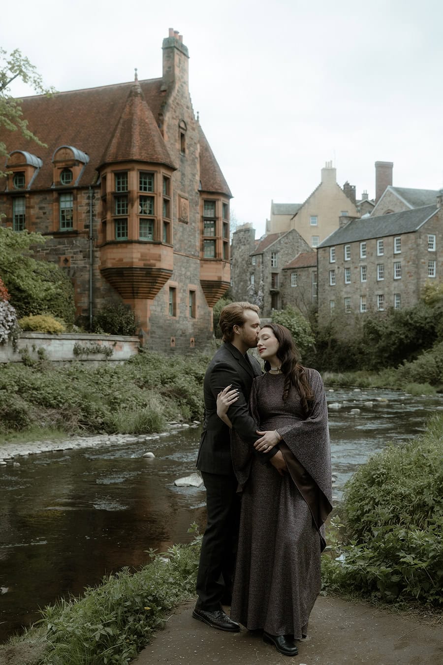 A man kisses his partner on the cheek as she closes her eyes and smiles, standing together beside the Water of Leith in Dean Village, Edinburgh, with Well Court rising behind them