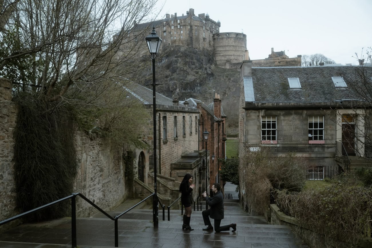 A man gets down on one knee to propose on the rain-soaked Vennel Steps in Edinburgh, with Edinburgh Castle rising dramatically on its volcanic rock above the Old Town rooftops behind them