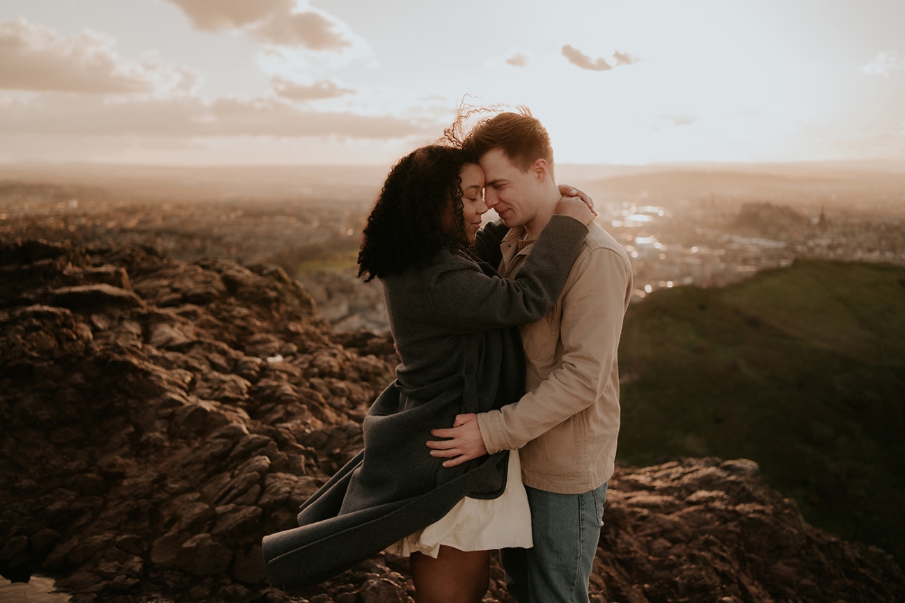 A couple embracing on a rocky hillside at sunset, overlooking a city below. The woman in a dark coat and light dress wraps her arms around the man in a beige jacket, their foreheads touching as golden light fills the sky