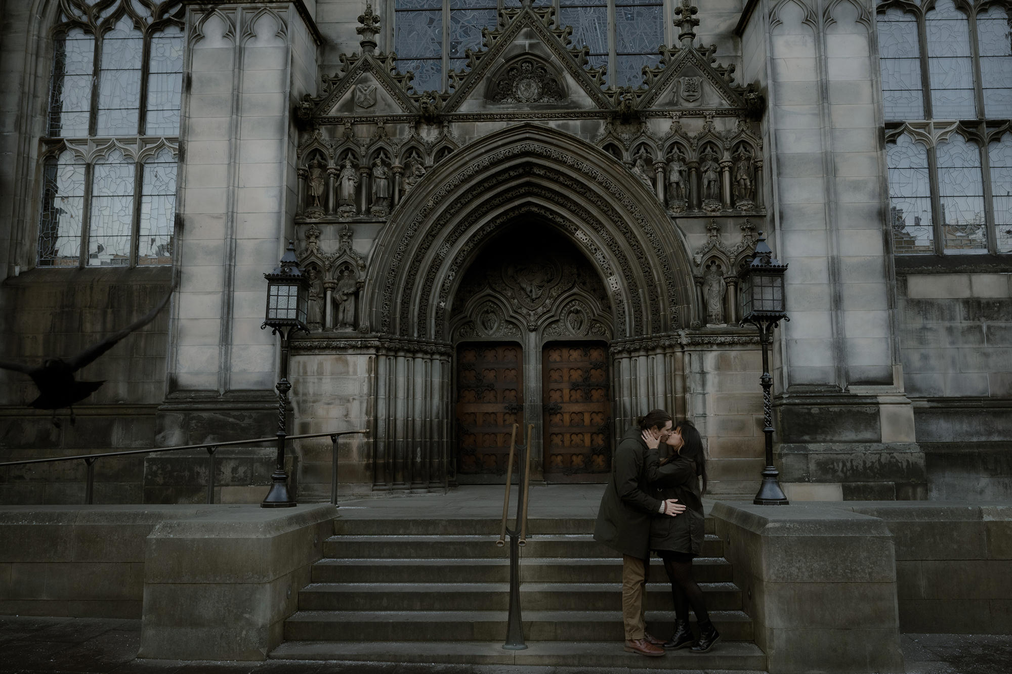 A couple shares a romantic kiss on the steps of a grand gothic-style St Giles Cathedral in Edinburgh Old Town. The intricate stone carvings, arched doorway, and large wooden doors serve as a backdrop, while a blackbird in flight adds a cinematic touch to the moody atmosphere