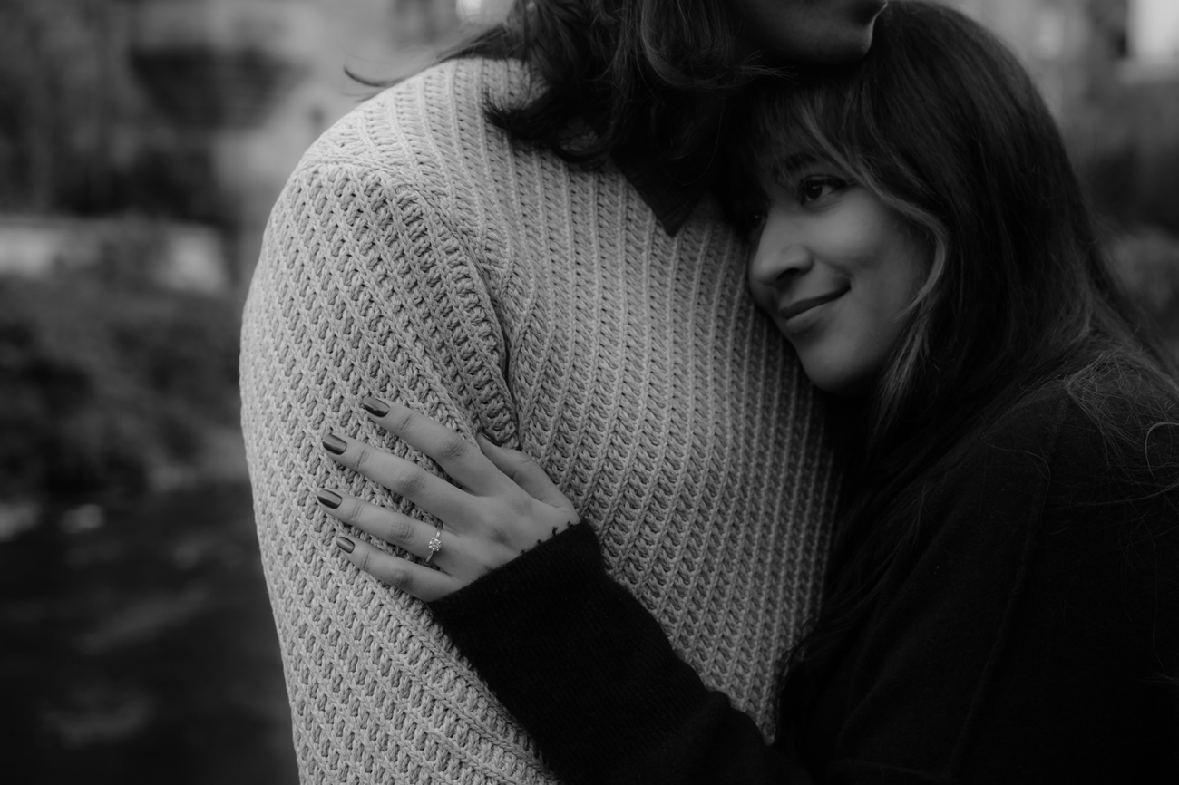 A close-up of a woman resting her head against her partner's chest, smiling softly. Her hand with an engagement ring on her finger, rests on his sweater