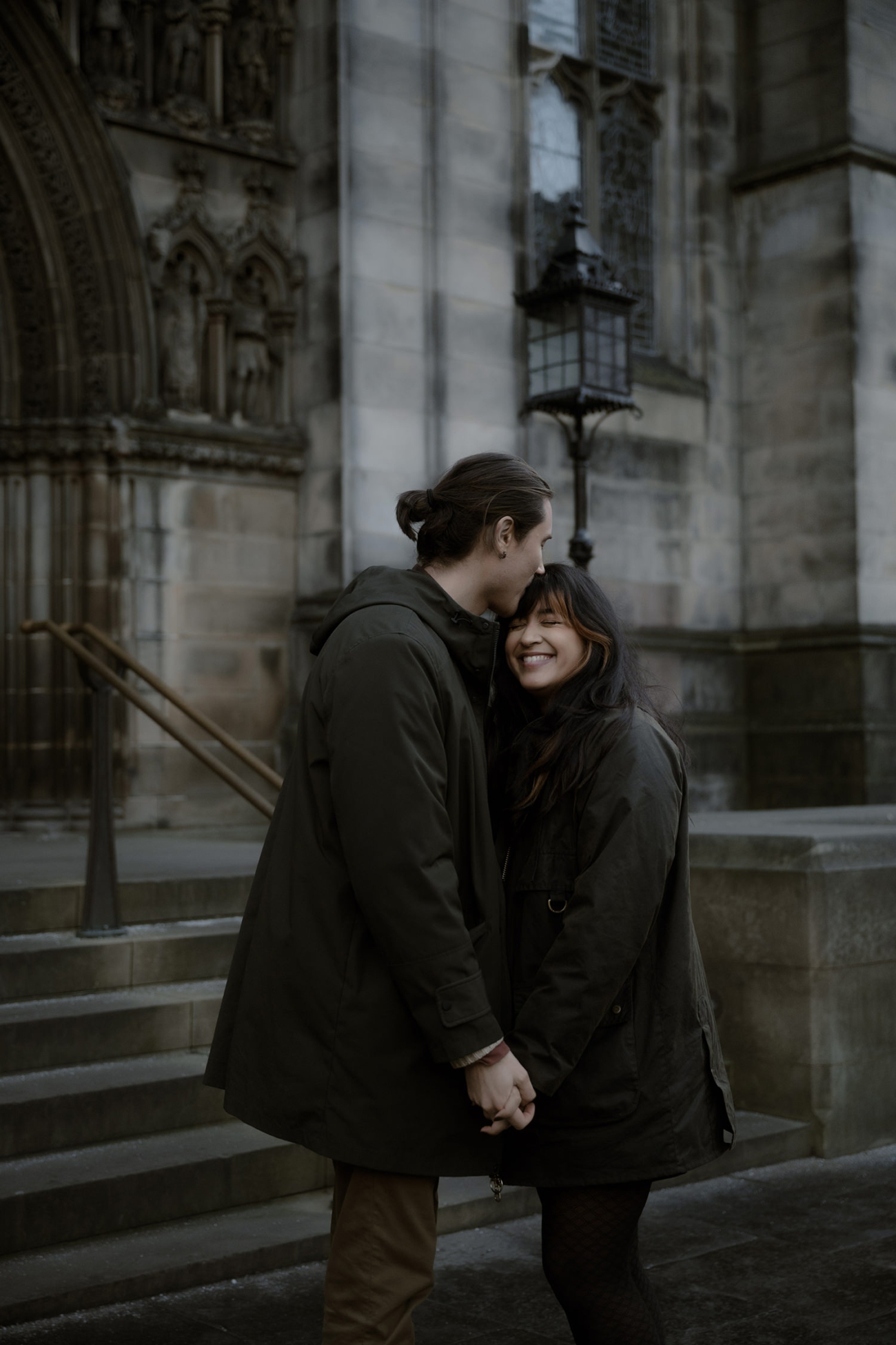 A couple holds hands in front of a historic stone church, gazing at each other with warm smiles. The man kisses the woman’s forehead