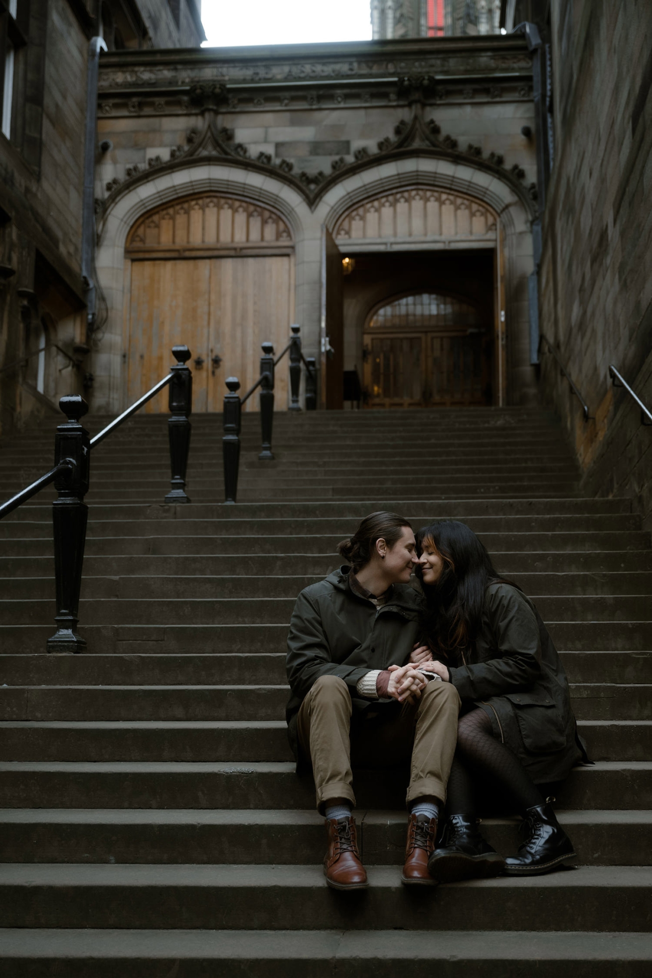 A couple sits on the steps of an old Edinburgh building, leaning in close with loving smiles