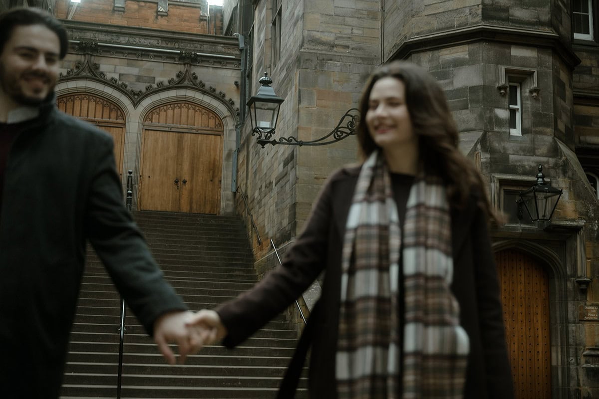 A couple hold hands and smile as they walk down the grand stone staircase of New College (Divinity College) in Edinburgh, the gothic arched doorway and Victorian gas lamps framing the scene behind them