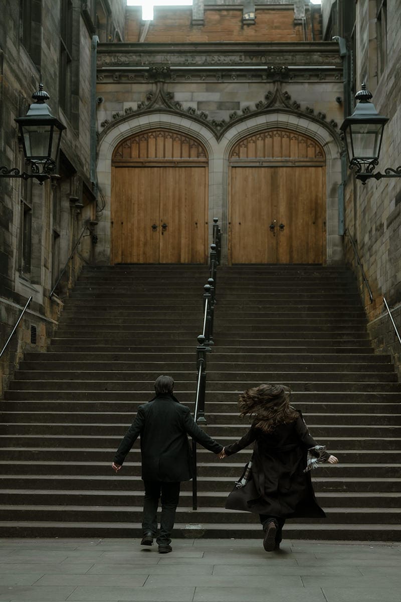 A couple run hand in hand up the sweeping stone staircase of New College (Divinity College) in Edinburgh, coats and scarves caught in the wind, the towering gothic double doors ahead of them