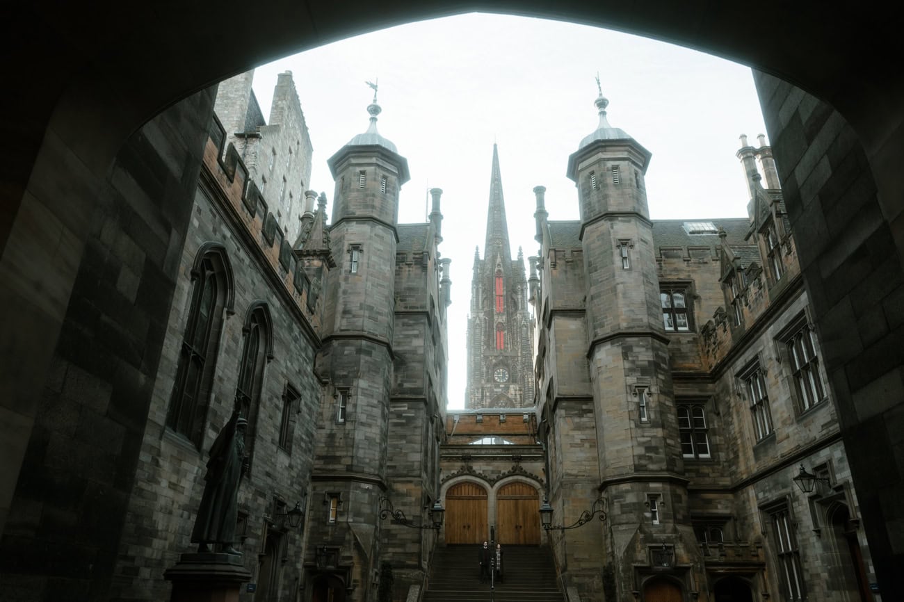 The dramatic gothic courtyard of New College (Divinity College) in Edinburgh, viewed through a stone archway, with turreted towers, grand stone staircases and the spire of the cathedral rising behind