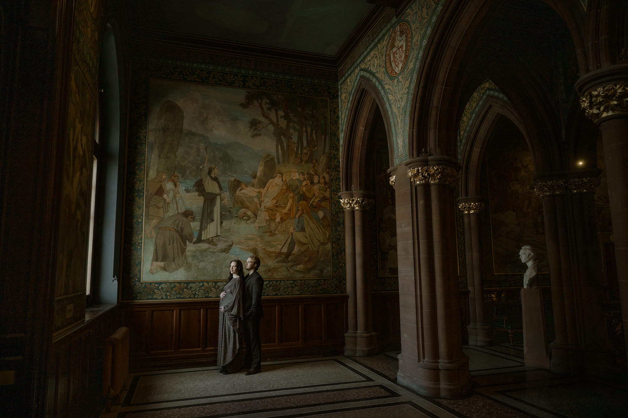 A couple stand together in the grand gallery of the Scottish National Portrait Gallery in Edinburgh, dwarfed by a vast historic mural, ornate gothic arches and gilded columns surrounding them