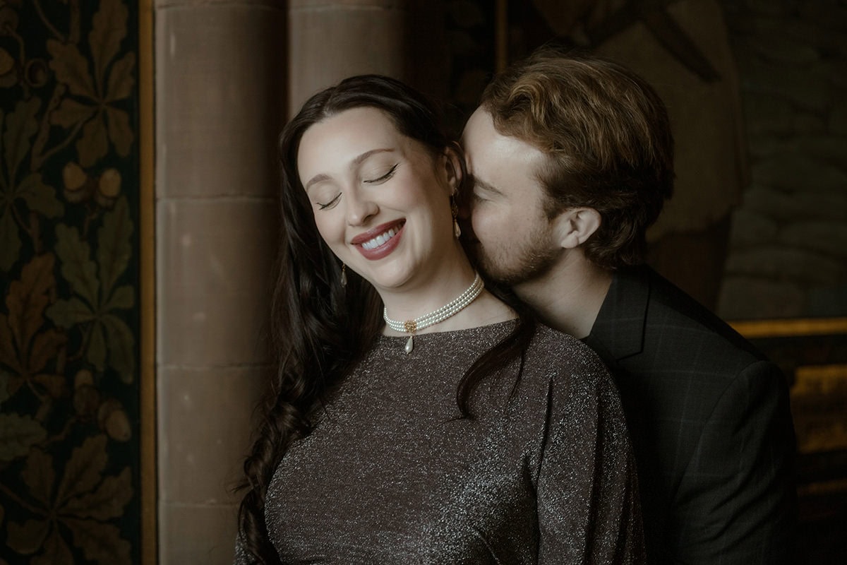 A man whispers to his partner from behind as she smiles with her eyes closed, photographed against the richly decorated interior of the Scottish National Portrait Gallery in Edinburgh
