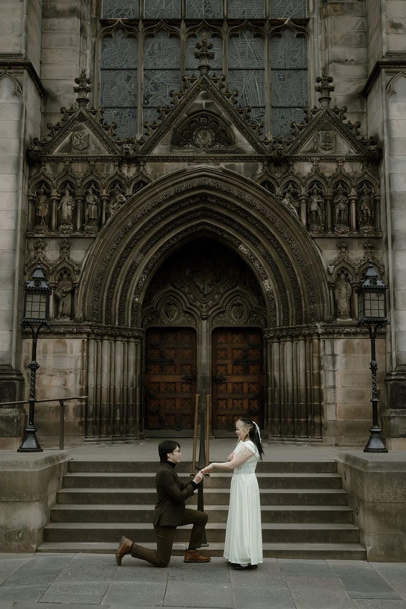 A man gets down on one knee to propose in front of the ornate gothic doorway of St Giles' Cathedral on the Royal Mile in Edinburgh, his partner in a white dress holding his hands