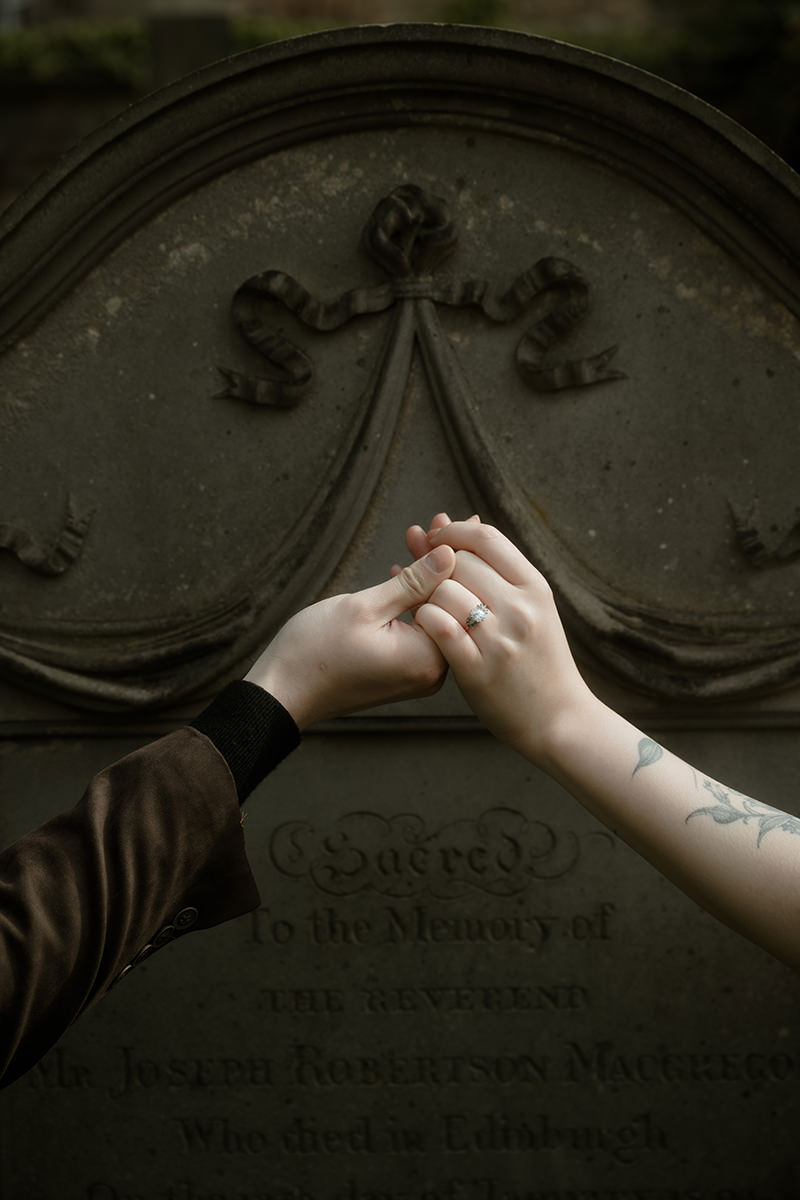 Close-up of an engaged couple holding hands against an ancient stone gravestone in Edinburgh, the woman's diamond engagement ring visible alongside a floral tattoo on her wrist