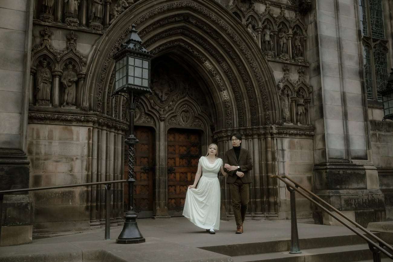 A couple walk arm in arm down the steps of St Giles' Cathedral in Edinburgh Old Town, the woman in a flowing white dress, framed by the cathedral's intricate gothic stone carvings and a Victorian lantern