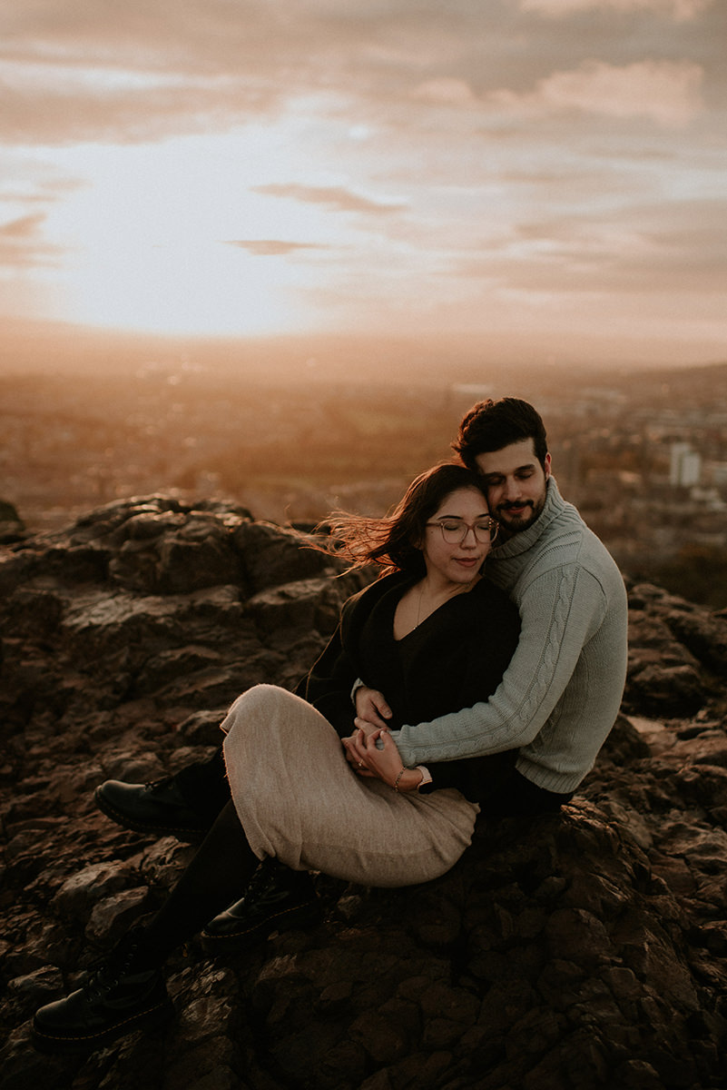 The couple seated on rocky terrain of Edinburgh Arthur's Seat at sunset, the man hugging the woman from behind as they look content and relaxed. The woman, wearing glasses and a light skirt with dark tights, leans into him while the city stretches out below in soft, golden evening light