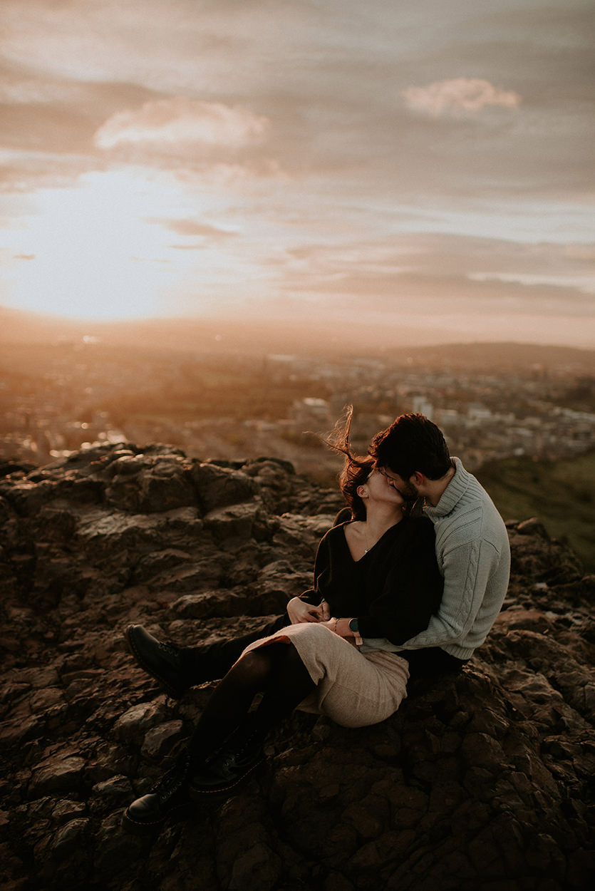 A couple sitting close together on a rocky hill at sunset, kissing softly as the wind lifts the woman’s hair. The golden sun glows low over the city in the background, casting warm light across the rugged landscape of Edinburgh