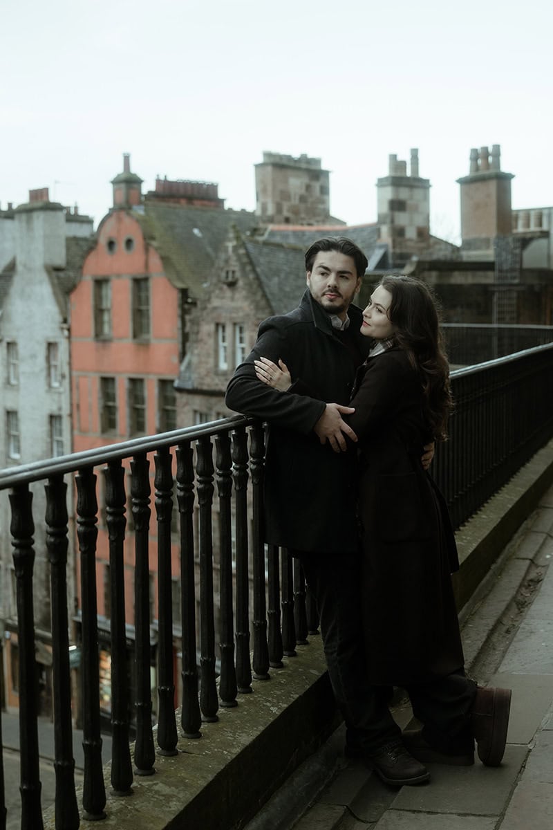 A couple embrace on the stone balcony of Victoria Terrace in Edinburgh, leaning against the iron railings with the colourful rooftops and historic tenements of the Old Town stretching behind them