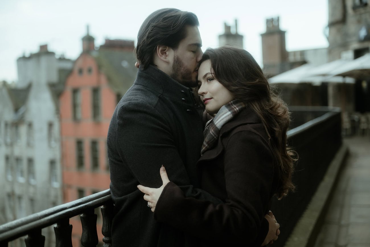 A man kisses his partner on the forehead as she smiles softly on Victoria Terrace in Edinburgh, the moody Old Town rooftops and a hint of Victoria Street below visible behind them