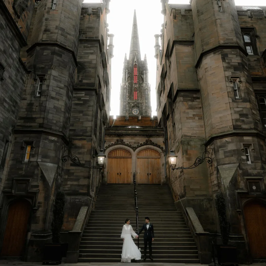 Romantic wedding photoshoot at The Divinity College (New College), Edinburgh - newlywed couple stands on the grand stone staircase of this historic Gothic building. The dramatic architecture and towering spire create a fairytale-like backdrop; Best Edinburgh Photography locations (Guide)