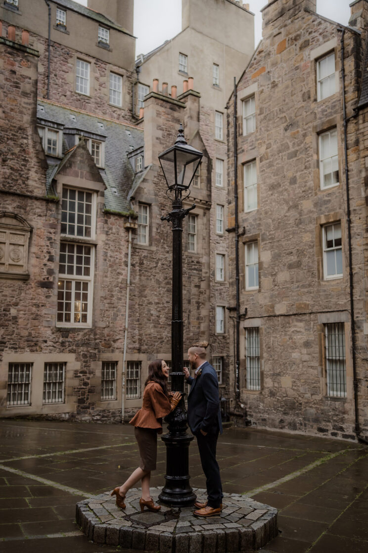 A couple playfully peek at one another around an old fashioned light post in Edinburgh during their Scotland elopement.