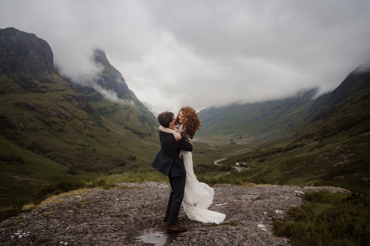 A groom lifts and spins his bride around at a scenic overlook during their Scotland elopement.