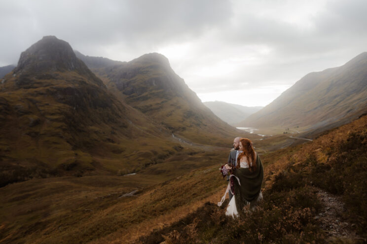 A bride wearing a tartan shawl embraces her husband after eloping in Scotland while they take in the view of a valley in Glencoe.
