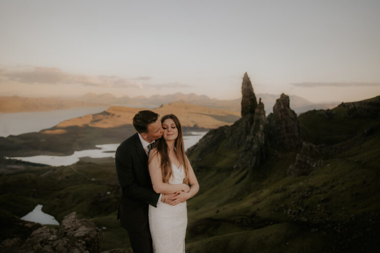 A groom embraces his new bride from behind in front of the Old Man of Storr rock formation during their Scotland elopement.