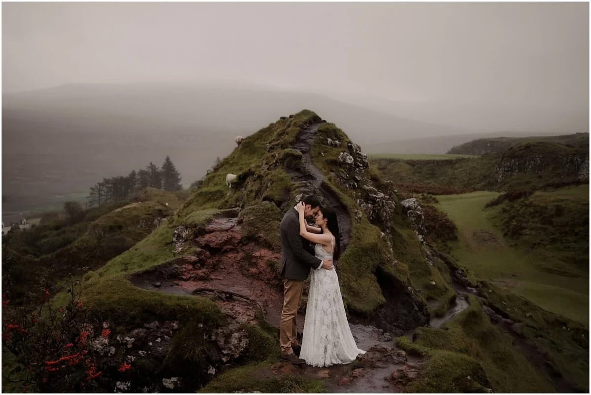 Couple embracing on the mossy red rock formation at Fairy Glen, Isle of Skye, with mist rolling over the hills behind them
