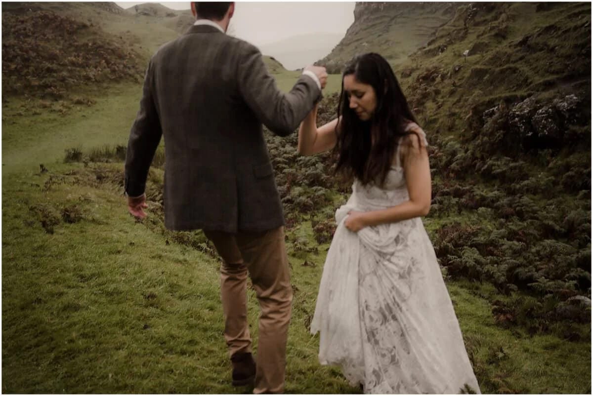 Groom helping his partner navigate the steep grassy path through Fairy Glen, Isle of Skye, in the rain