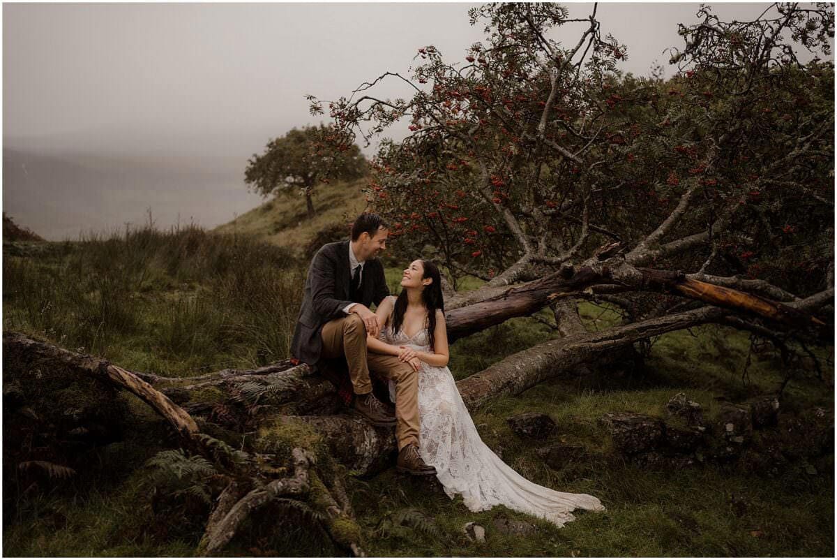 Couple sitting together on a fallen rowan tree at Fairy Glen, Isle of Skye, smiling at each other on a misty autumn day