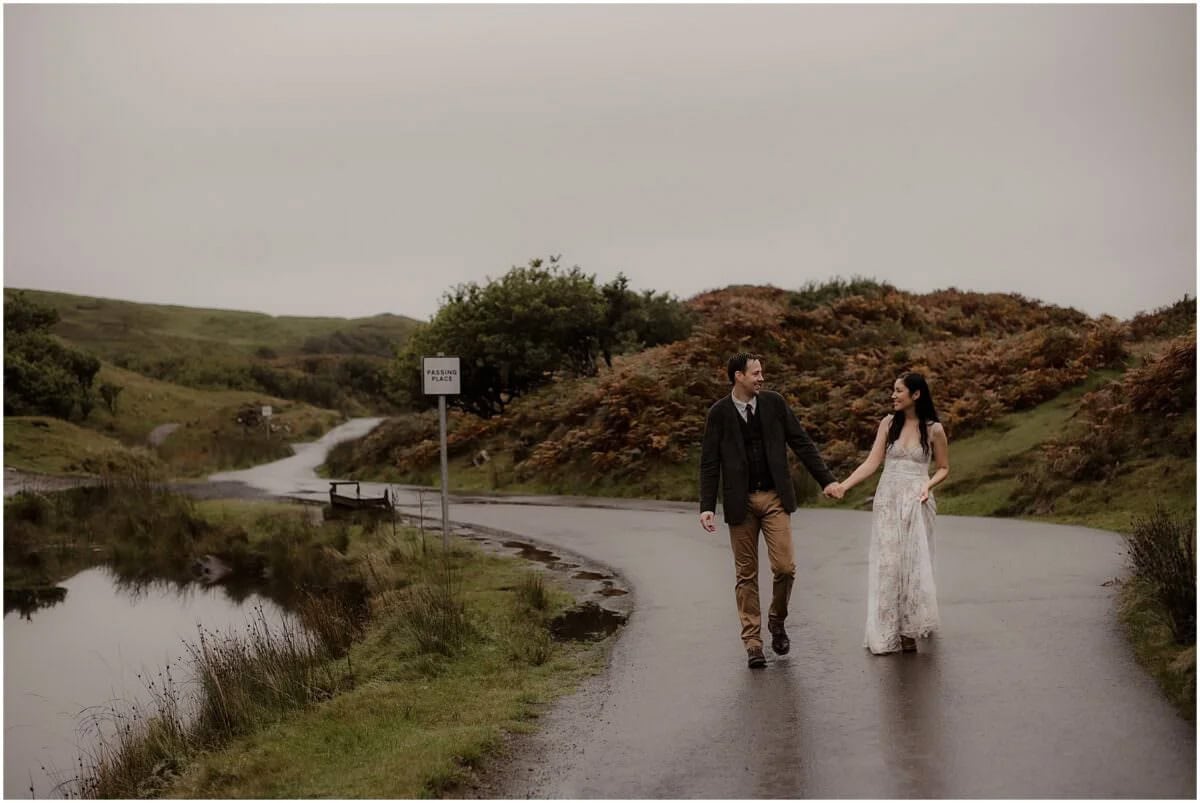 Couple walking hand in hand along a single-track road near Fairy Glen, Isle of Skye, passing a passing place sign in the rain
