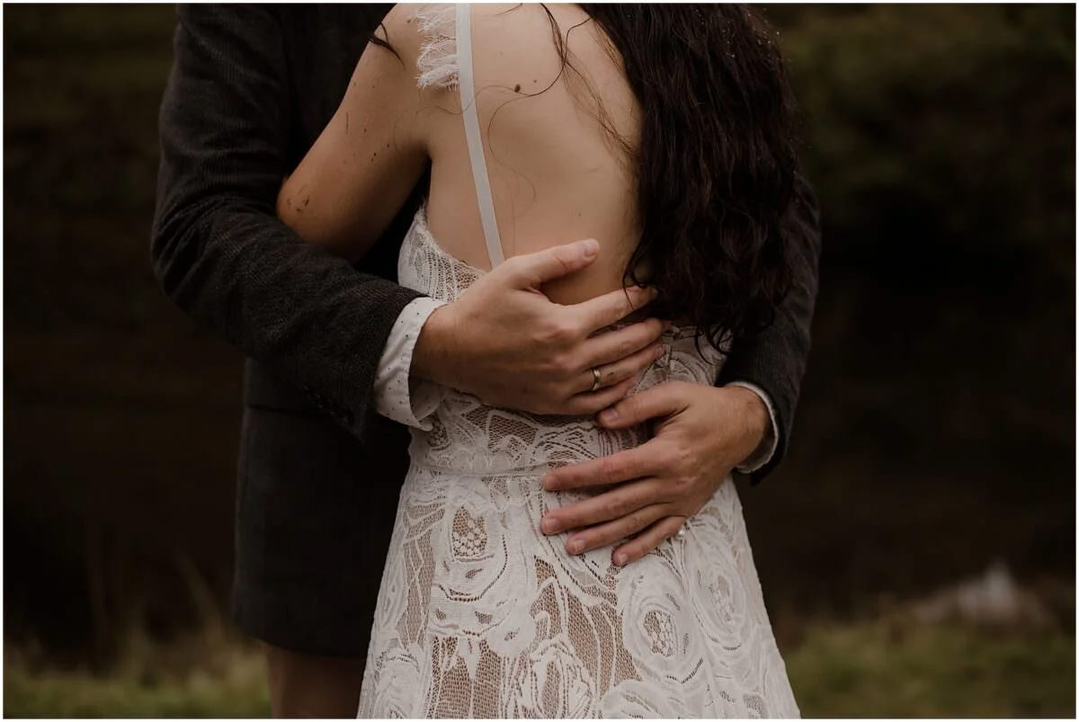 Close-up of a groom's hands around his partner's waist, lace wedding dress detail visible, at Fairy Glen Isle of Skye