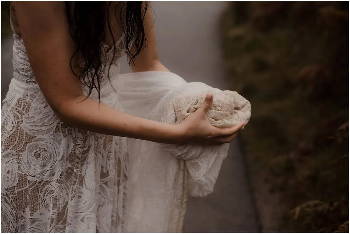 Bride gathering her wet lace wedding dress train in her hands during a rainy elopement at Fairy Glen, Isle of Skye