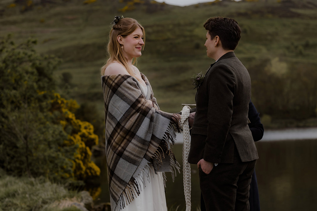 Bride wrapped in a tartan shawl and her partner bride in a dark tweed suit hold a woven handfasting cord as they face each other on the grassy shore of Dunsapie Loch under the rolling green hills of Holyrood Park