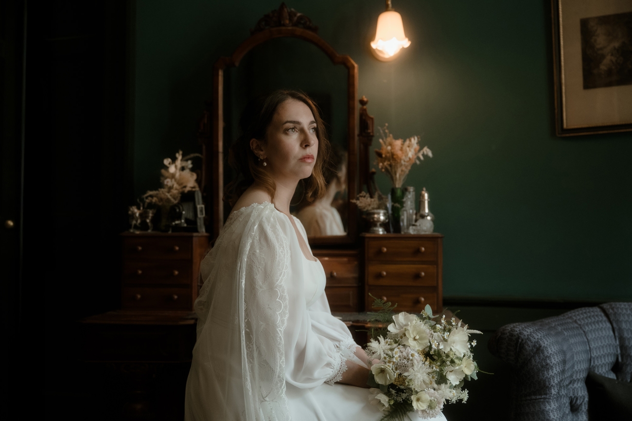 Bride in a flowing white gown with sheer lace bishop sleeves sits at an antique wooden vanity in a dark green–walled bridal suite, holding a cream-and-pale-yellow bouquet beside silver and glass dressing accessories