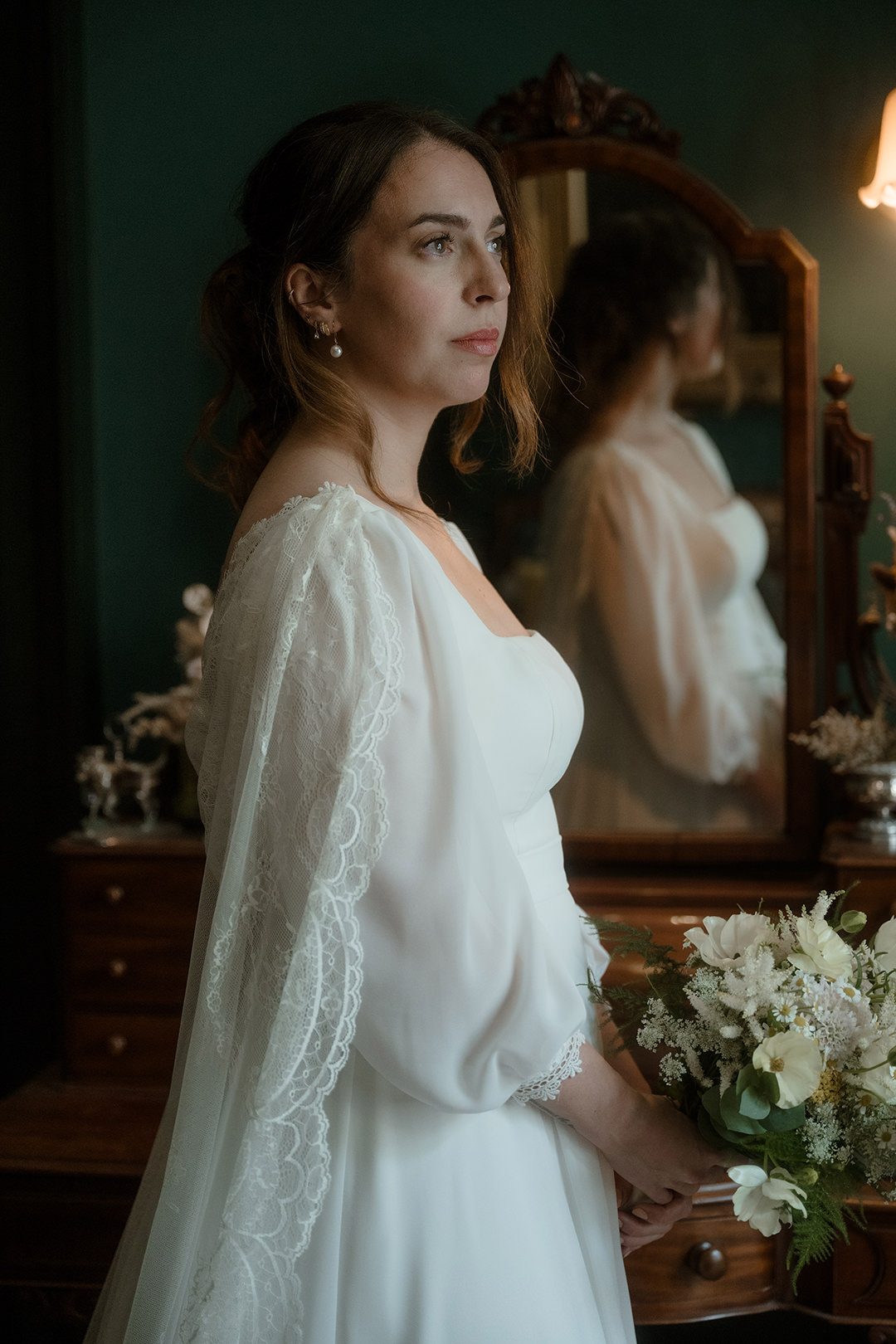 Close-up portrait of a bride in a square-neck wedding dress with scalloped lace sleeves, standing before an ornately carved mirror on a dark green wall and clutching a delicate ivory bouquet