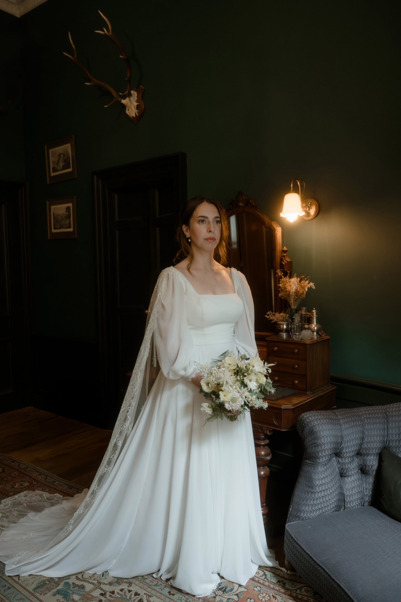 Full-length shot of a bride in a vintage-inspired long-sleeve gown and lace-edged veil holding a wild-flower bouquet in a wood-paneled room with dark green walls and an antique dressing table