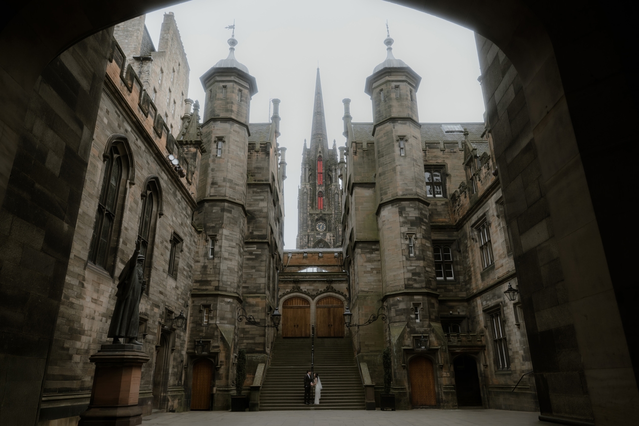 Bride in a flowing white gown and groom in a dark suit stand together at the foot of a grand stone staircase in a narrow medieval courtyard, framed by an arched gateway and twin towered walls, with a soaring Gothic spire and glowing red stained-glass windows rising above them on their Edinburgh wedding day