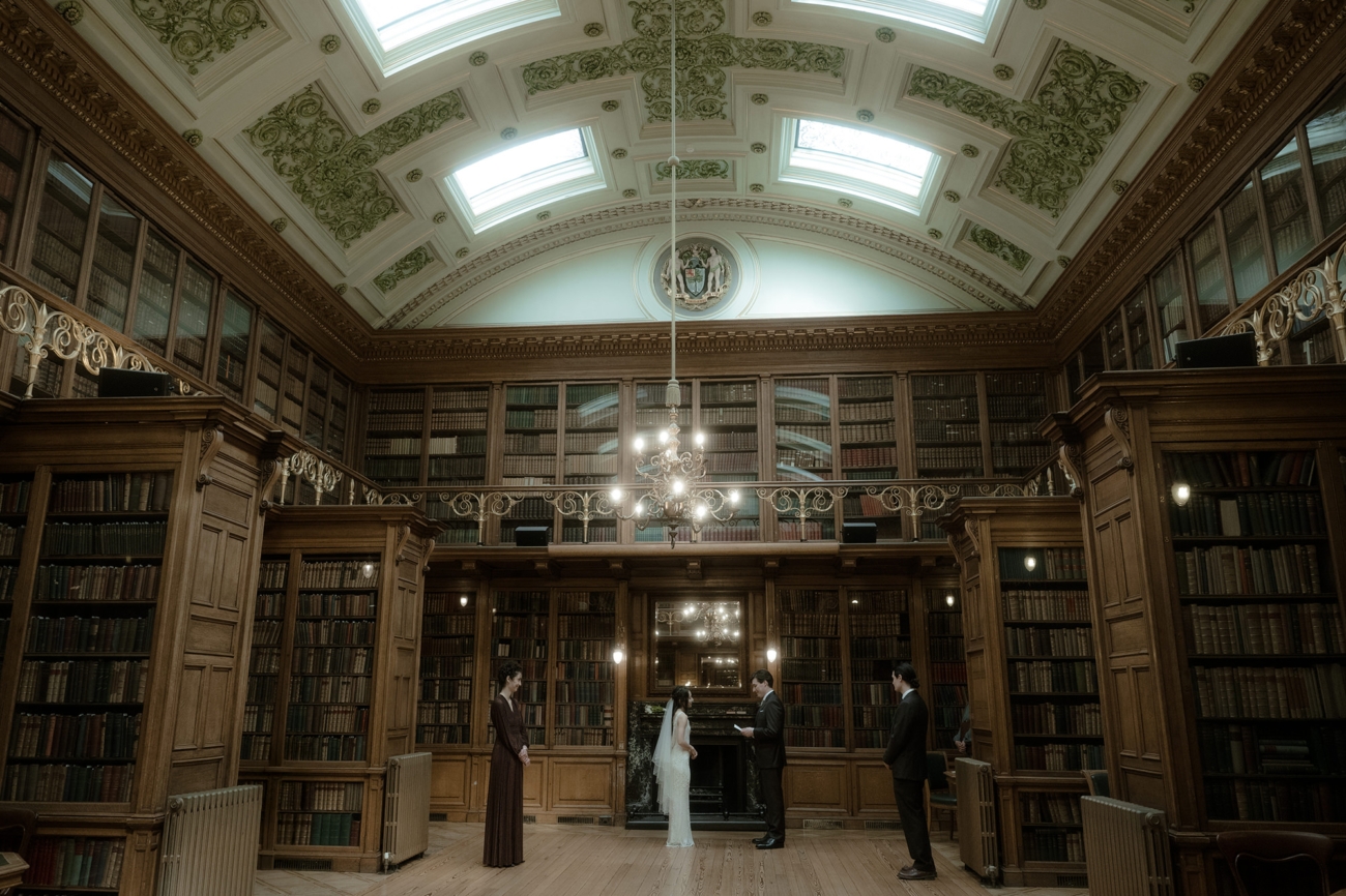 Bride and groom exchange vows in the Royal College of Physicians of Edinburgh library, surrounded by maid of honour and best man standing by the wooden shelves